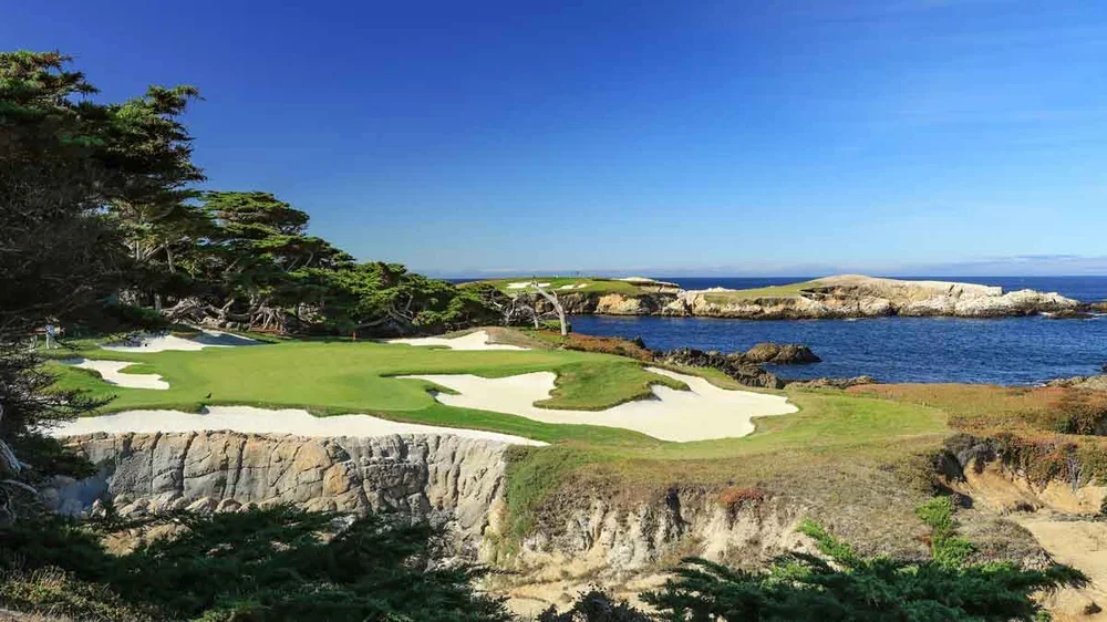 A golf course at Cypress Point with a green and white sand bunkers overlooks the ocean, surrounded by rocky cliffs, trees, and blue sky on a sunny day.